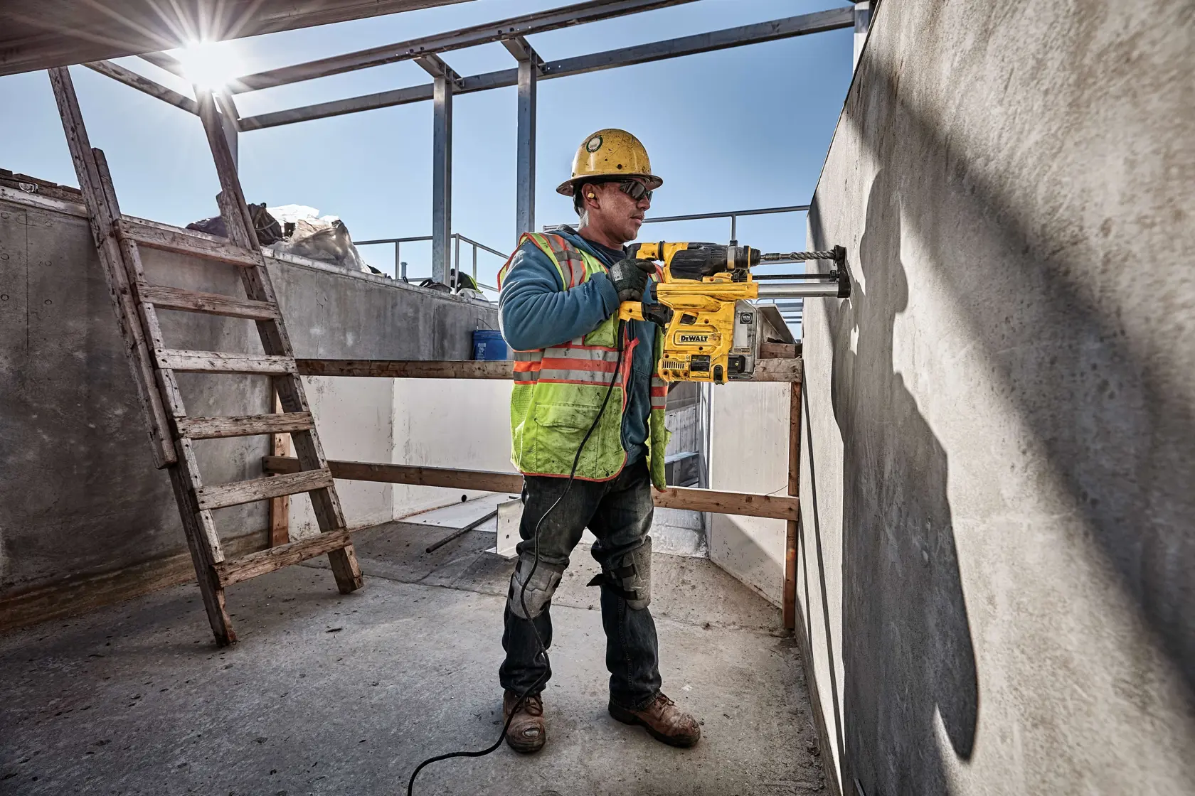 Ein Bauarbeiter mit Helm und Sicherheitsweste verwendet einen DEWALT D25333K Schlagbohrhammer, um eine Betonwand auf einer Baustelle im Freien zu bohren.