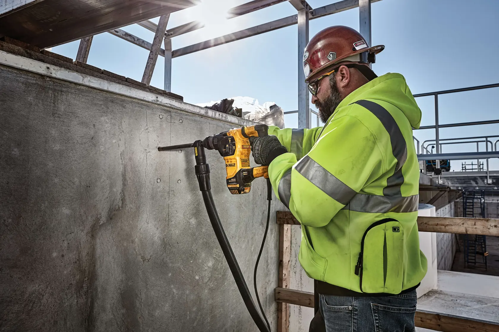 Ein Bauarbeiter mit Sicherheitsweste, Helm und Handschuhen bedient einen DEWALT-Bohrhammer (Modell D25333K), um eine Betonwand auf einer Baustelle im Freien zu bohren, umgeben von Metallträgern und Bauelementen. Das Gesicht des Arbeiters ist absichtlich verschwommen.
