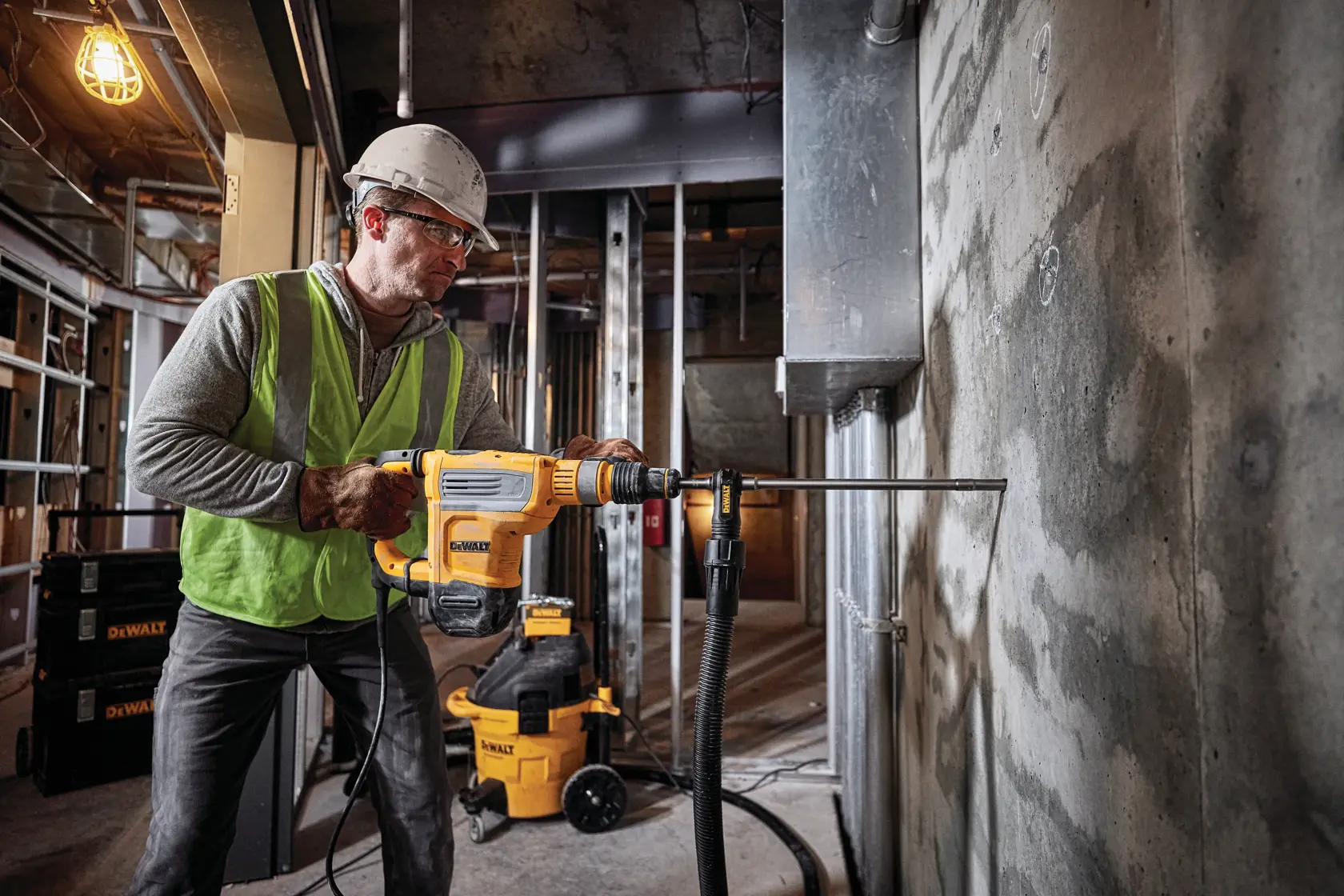 Ein Bauarbeiter mit Sicherheitshelm und Weste verwendet einen gelben DEWALT D25614K Abbruchhammer mit Staubabsaugung an einer Ziegelmauer.