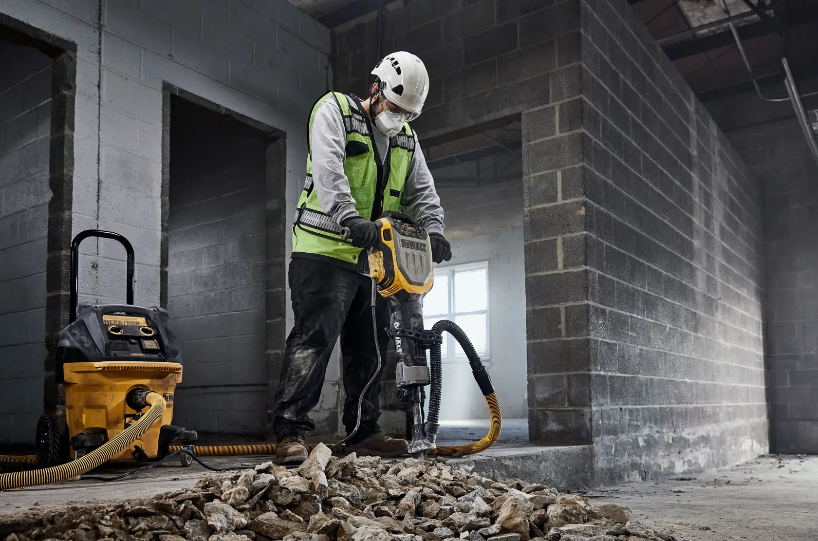 Ein Bauarbeiter mit Sicherheitsweste und Helm benutzt einen DEWALT Presslufthammer (Modell D25966), um Beton in einem unfertigen Gebäude zu zertrümmern. Ein DEWALT Staubabsauger ist an den Presslufthammer angeschlossen, und zerbrochene Betonstücke liegen auf dem Boden.
