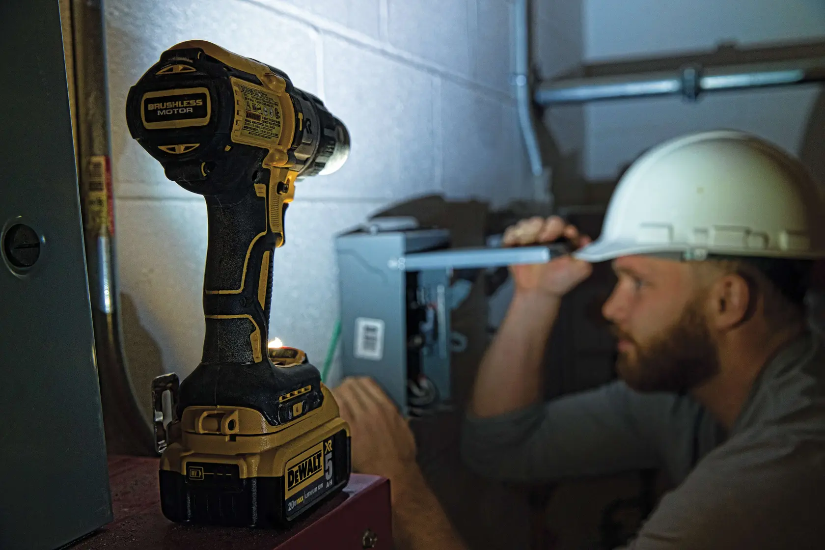 Eine Person mit einer Warnjacke und einem Bauhelm benutzt einen DEWALT Akku-Bohrschrauber (Modell DCD996P2), um in eine Beton- oder Mauerwerkswand in einer Industrie- oder Baustellenumgebung zu bohren. Das Produkt und der Griff am Werkzeug sind deutlich zu sehen.