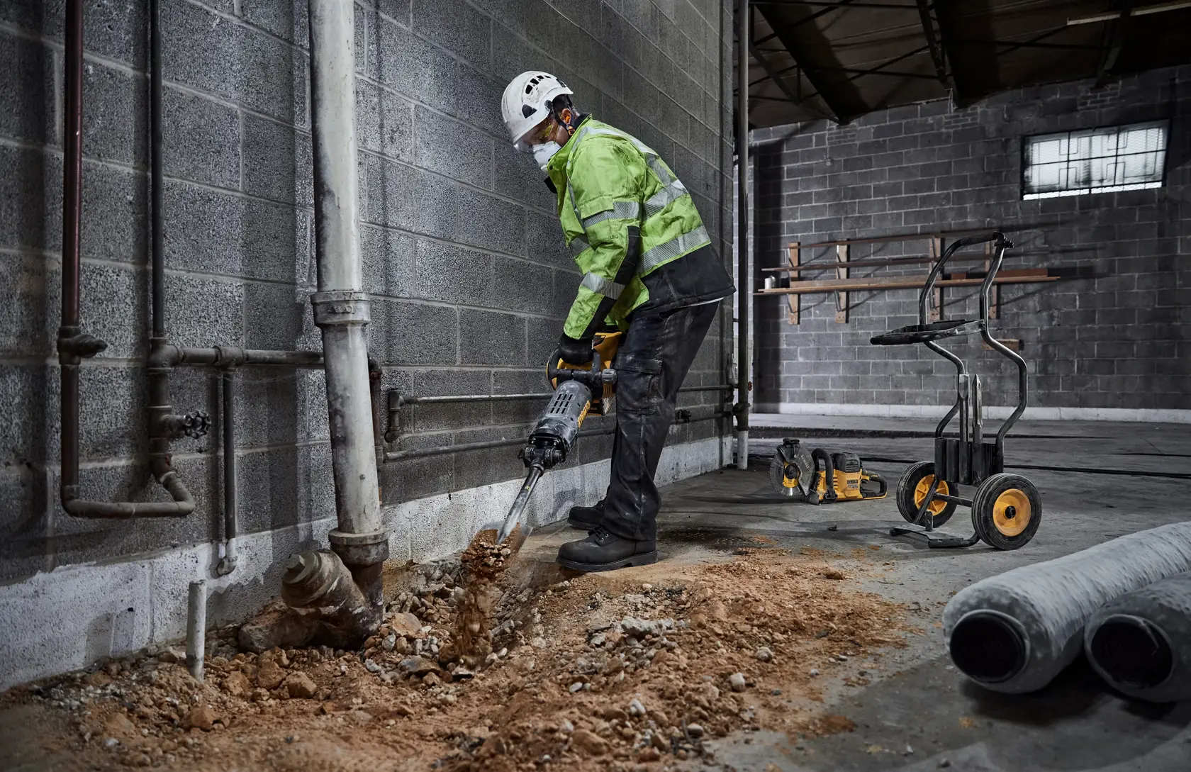 A person wearing safety gear uses a DEWALT demolition hammer to break up a concrete floor near a wall in an industrial setting. Construction tools and equipment are visible in the background.