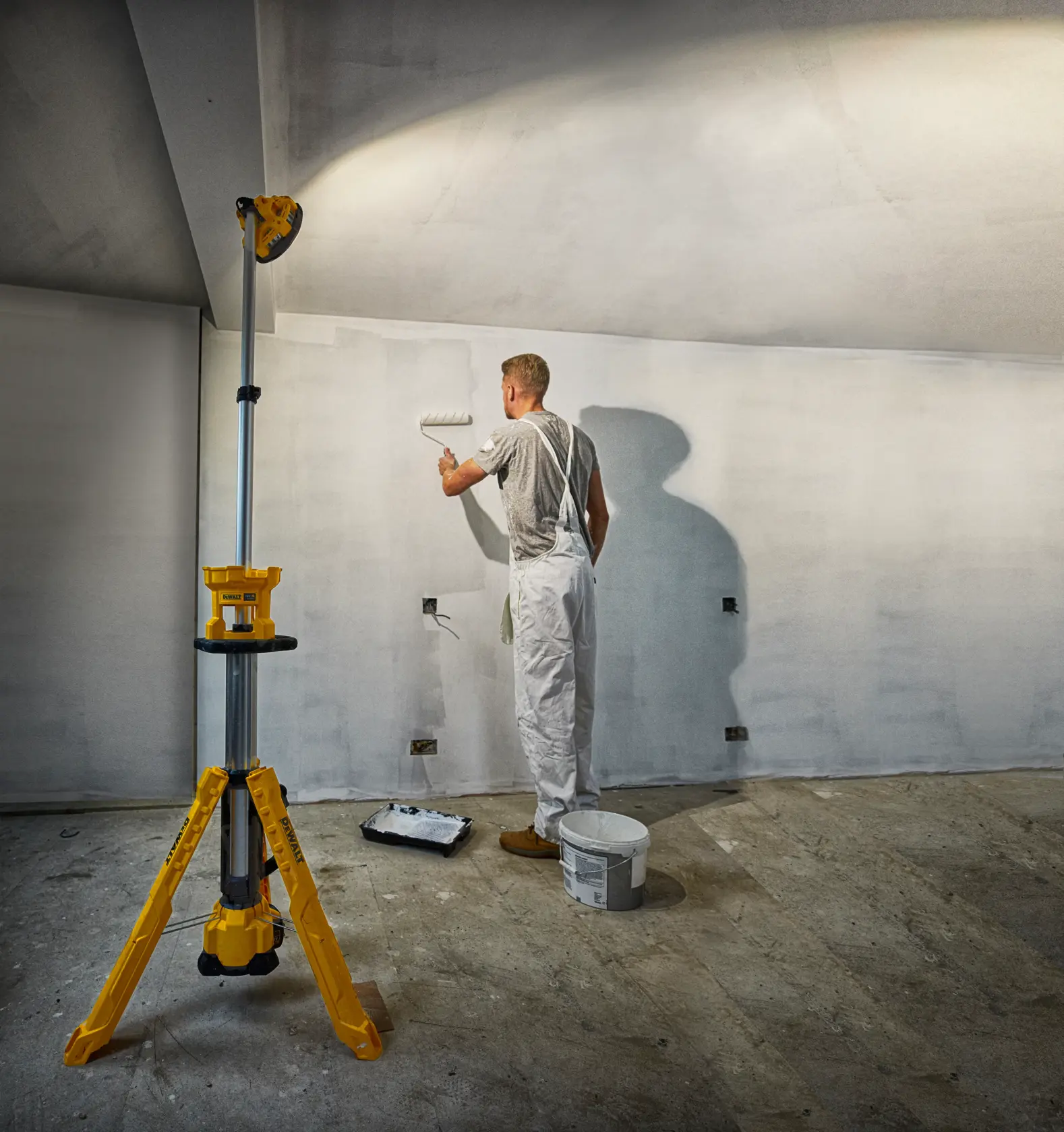Ein Maler arbeitet mit einer Farbrolle, beleuchtet von einem gelben DEWALT Stativ-Arbeitslicht im Renovierungsraum.