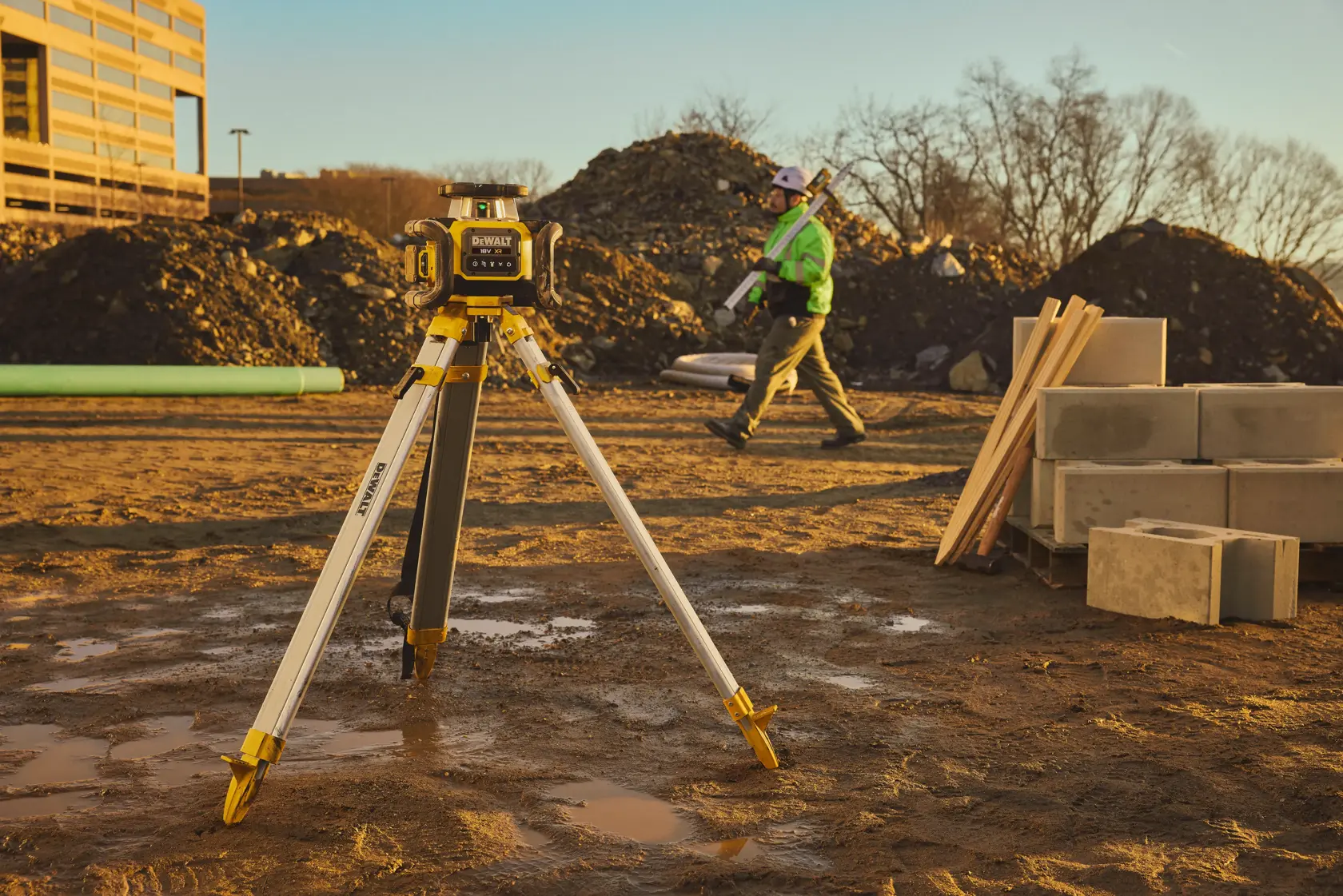 DEWALT Laser-Nivelliergerät auf Stativ auf schlammiger Baustelle bei Sonnenuntergang. Arbeiter und Materialien.