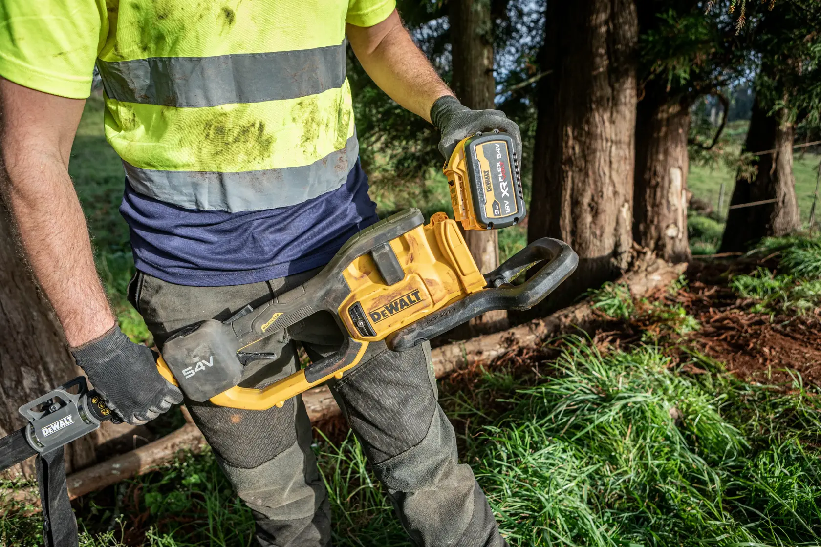 Person mit einer DEWALT Akku-Kettensäge im Freien, trägt Arbeitshandschuhe und Warnshirt.
