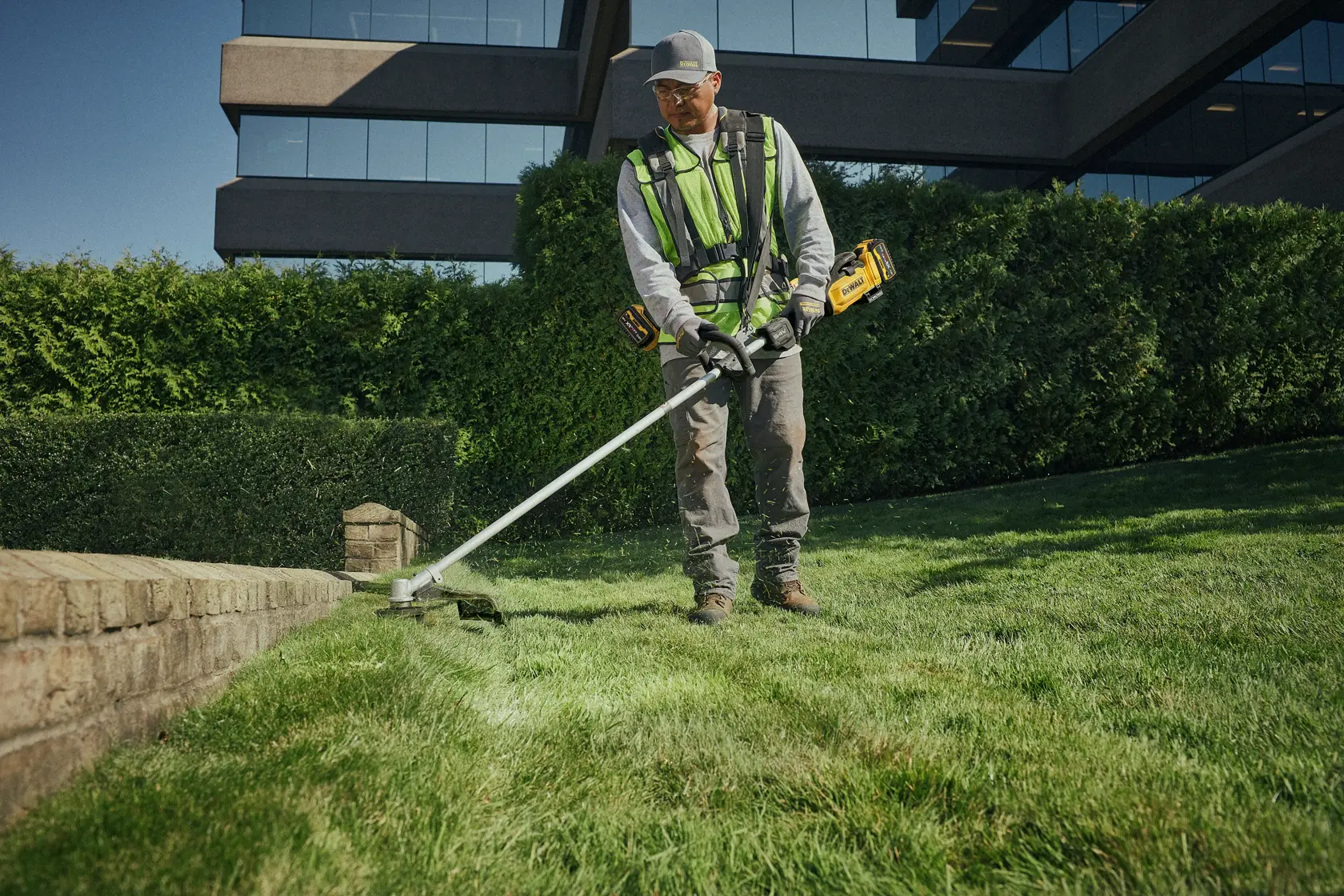 Person trimmt Gras mit einem DEWALT DCMST901N Akku-Trimmer an einer gemauerten Kante im Garten.