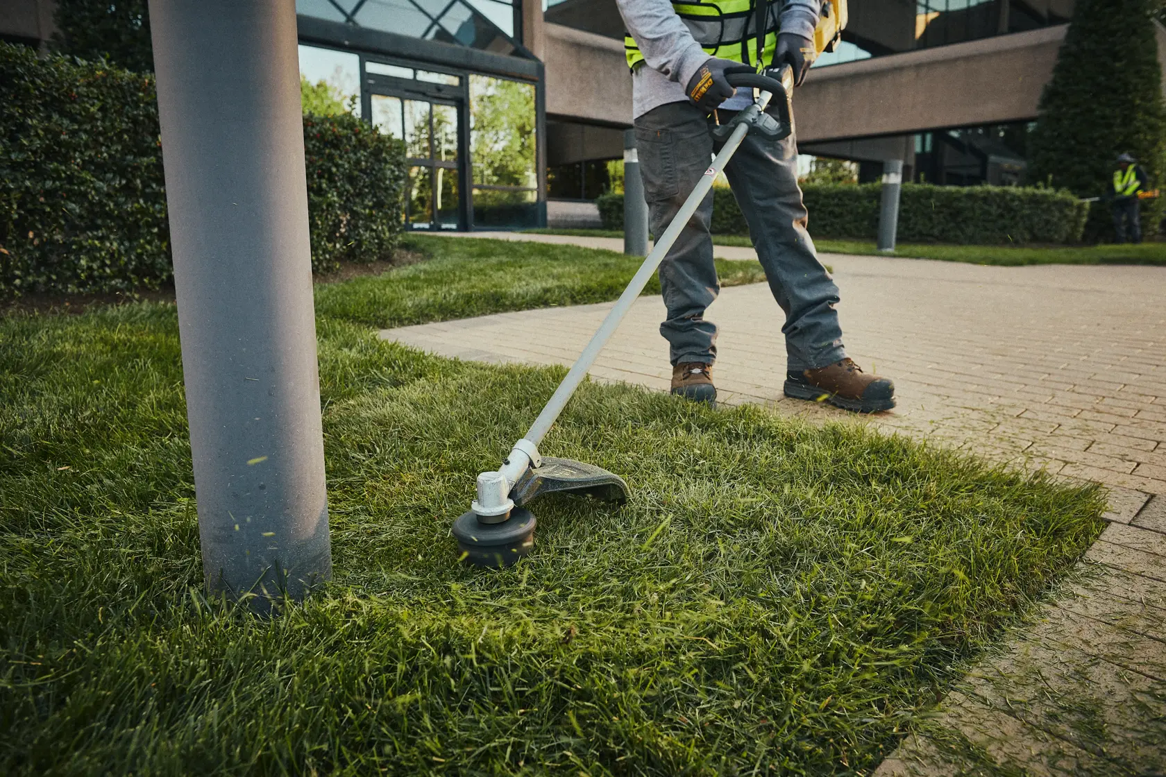 Person trimmt Gras mit kabellosem DEWALT-Trimmer neben Gehweg und Laternenmast im Außenbereich.
