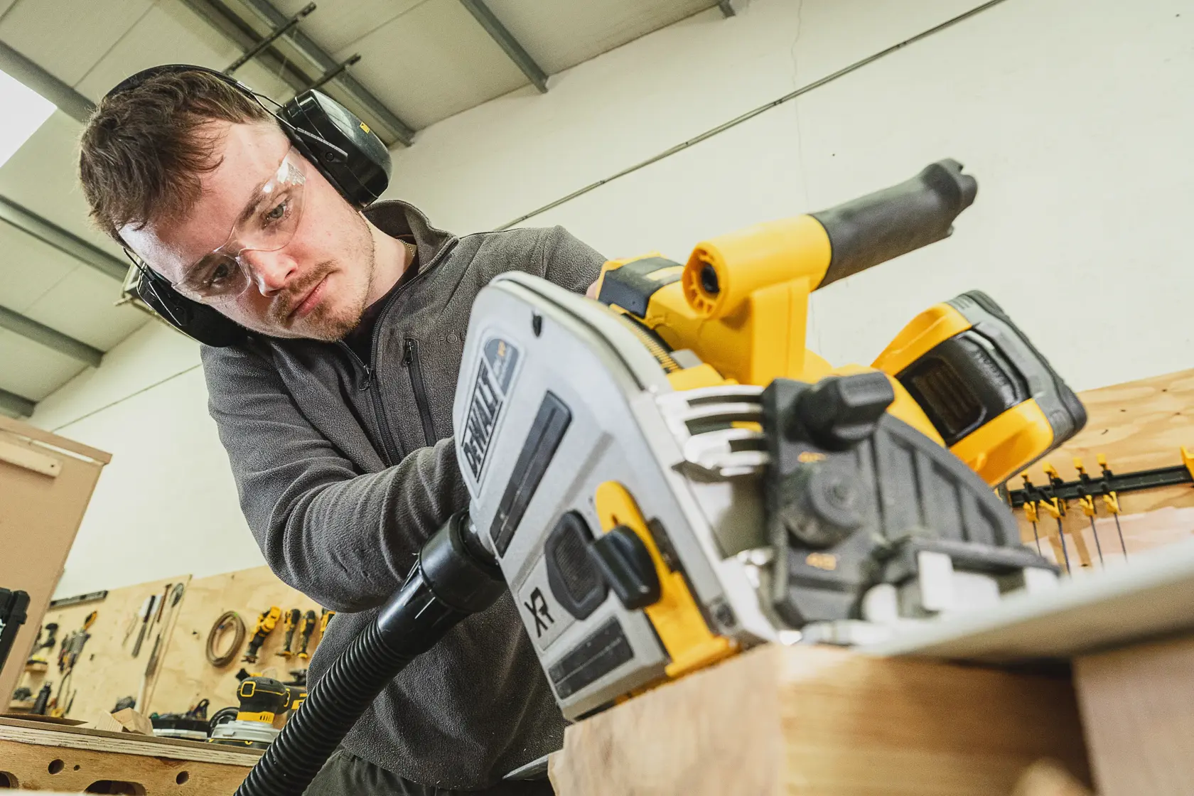Person sägt Holz mit einer DEWALT Akku-Säge, trägt Schutzbrille und Gehörschutz in einer Werkstatt.
