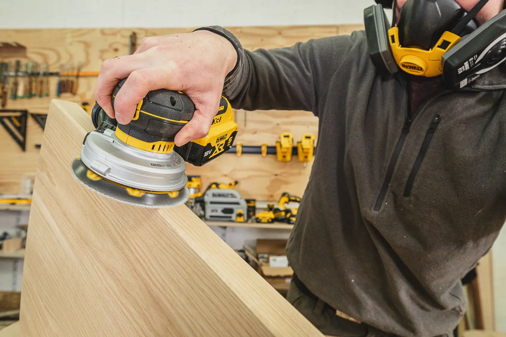 Person schleift Holz mit einem DEWALT Akku-Schleifer in einer Werkstatt und trägt eine Schutzmaske.