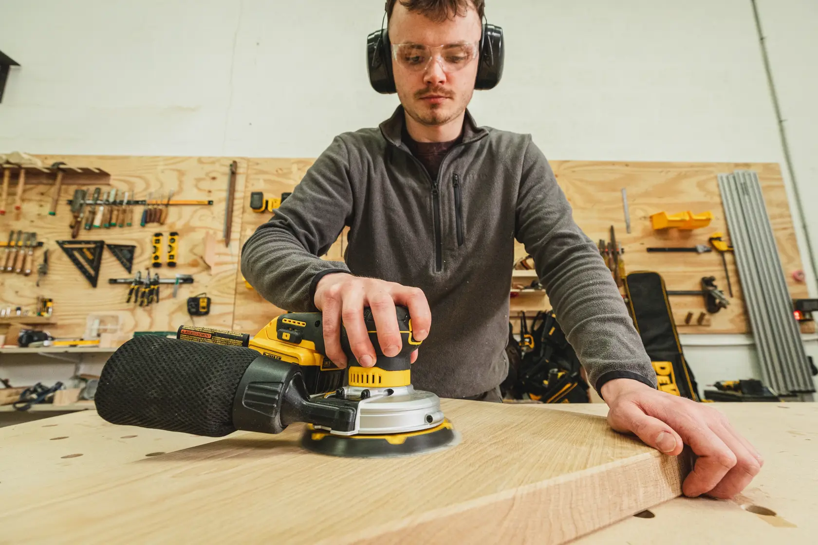 Person schleift ein Holzbrett mit einem DEWALT-Gerät in einer Werkstatt mit Werkzeugen im Hintergrund.