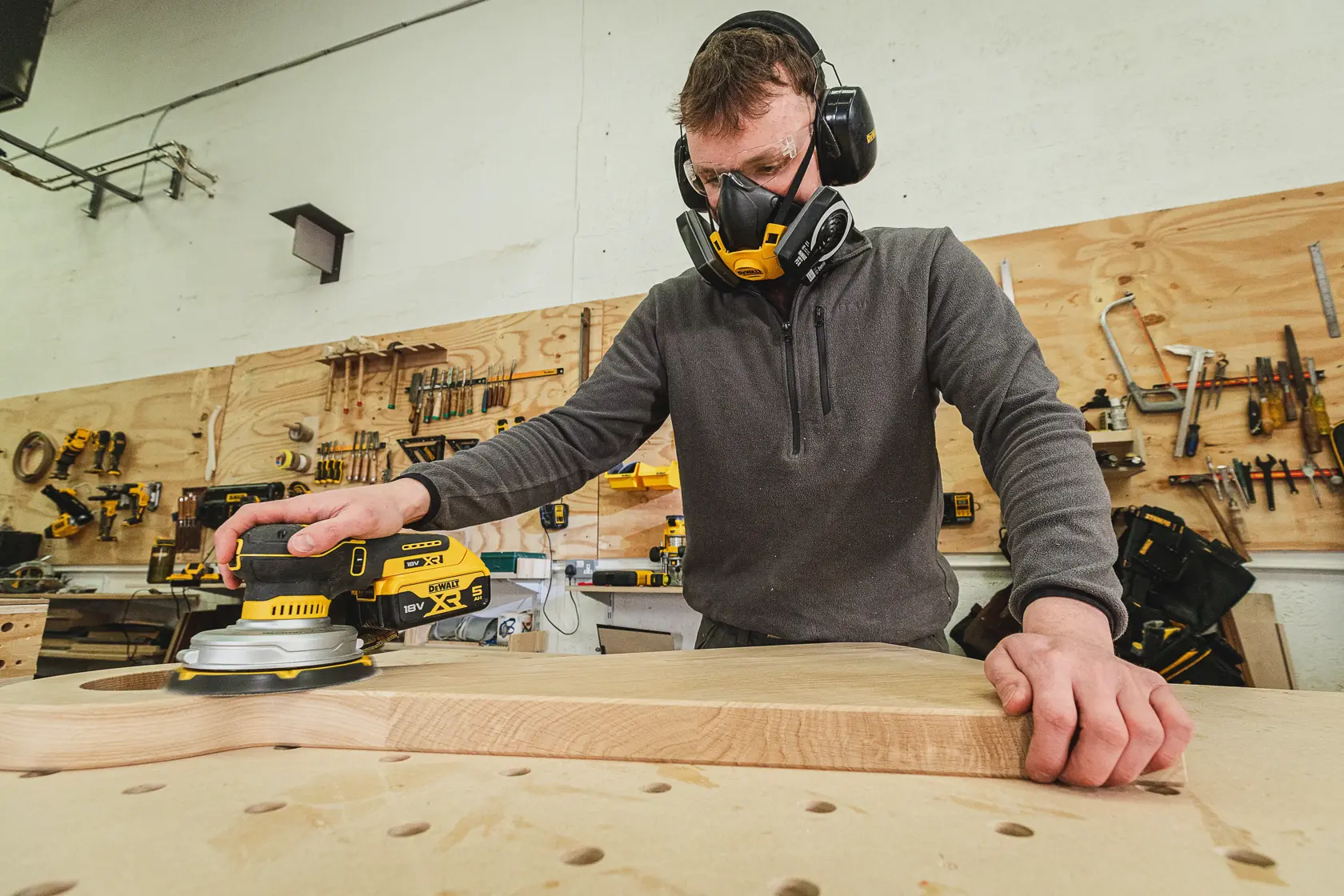 Person schleift Holz mit DEWALT Schleifer, trägt Schutzkleidung in einer Werkstatt mit Werkzeugen an der Wand.