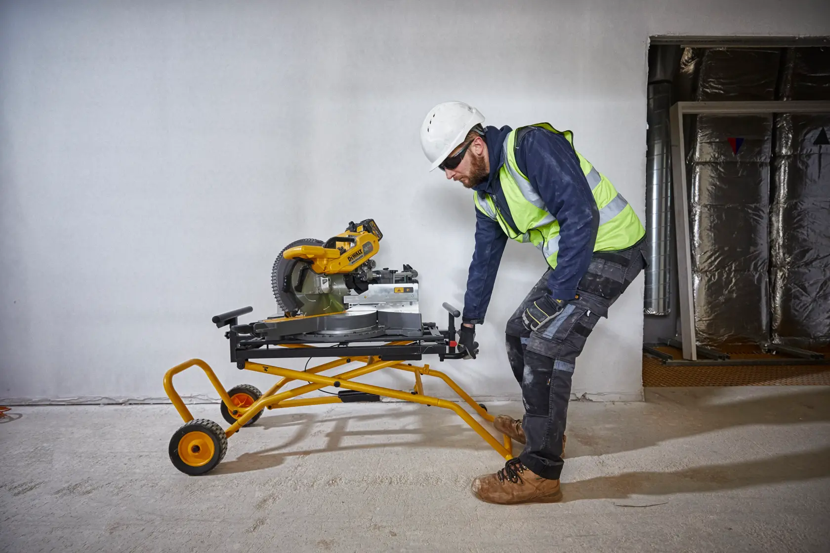 Arbeiter bewegt eine DEWALT Kappsäge auf gelbem Rollgestell in einer Baustelle.