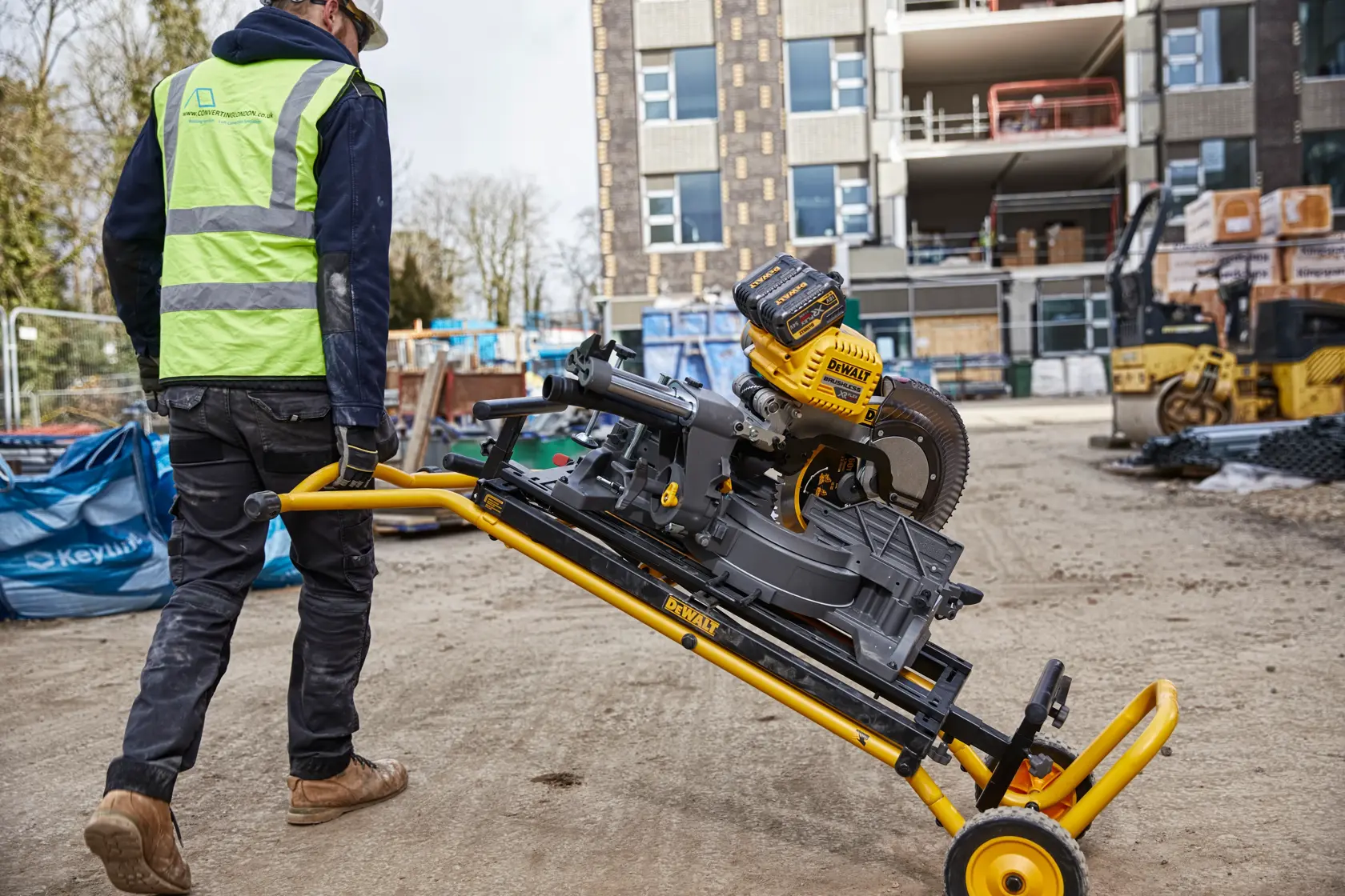 Arbeiter bewegt eine DEWALT Kappsäge auf einem Rollgestell auf einer Baustelle mit Gebäuden im Hintergrund.