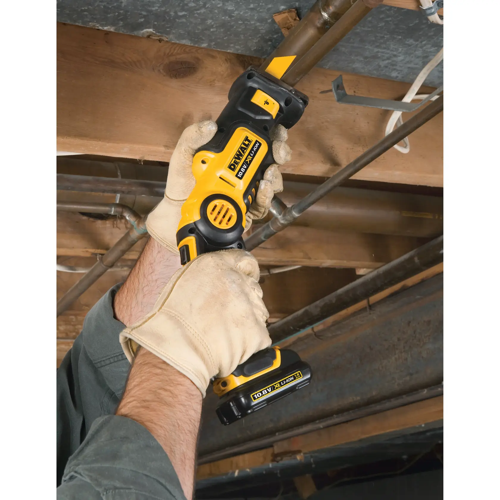 Gloved hands using a DEWALT cordless reciprocating saw to cut a pipe in an unfinished ceiling area.