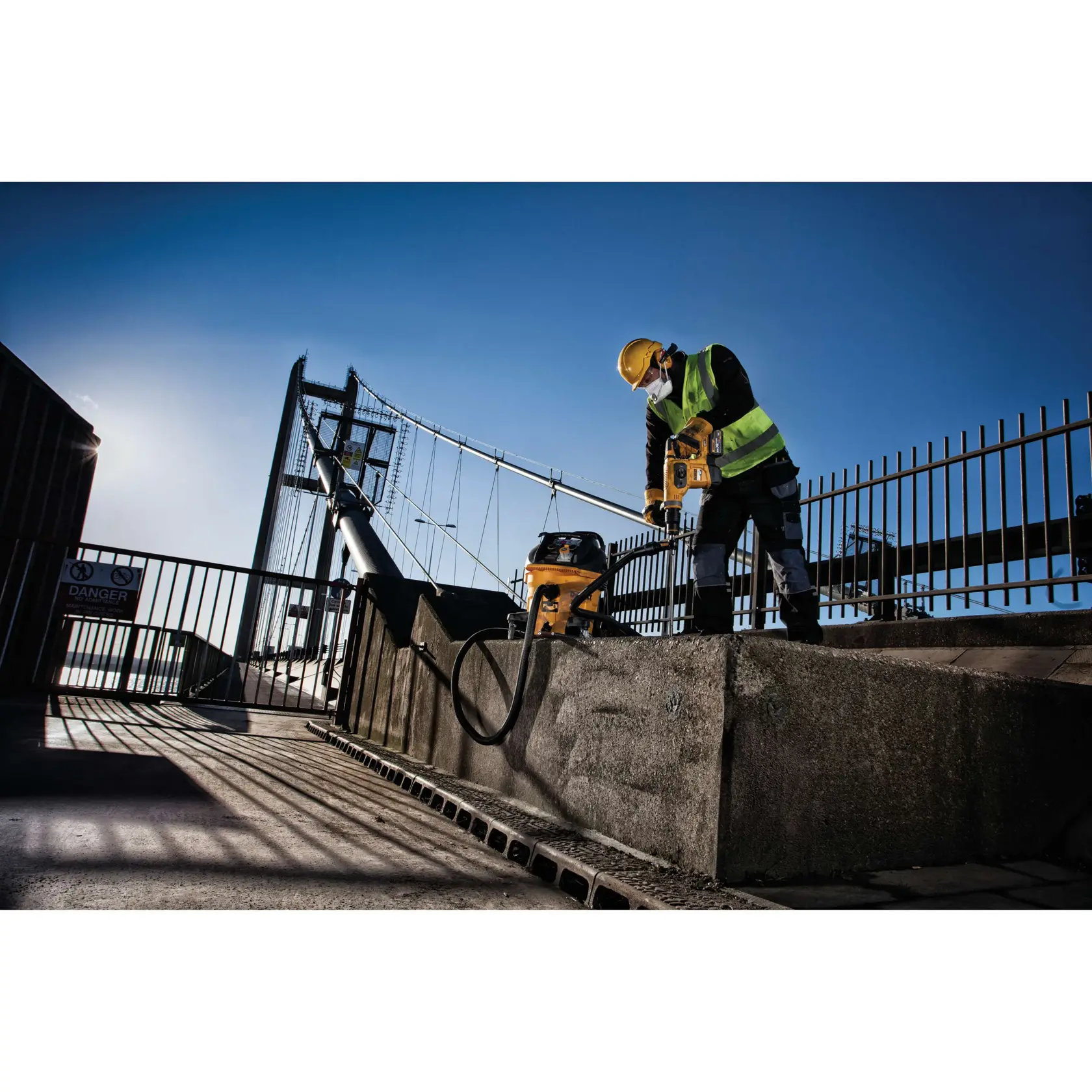 Trabajador usa herramienta DEWALT y aspiradora en cemento cerca de puente, con chaleco y equipo de seguridad.