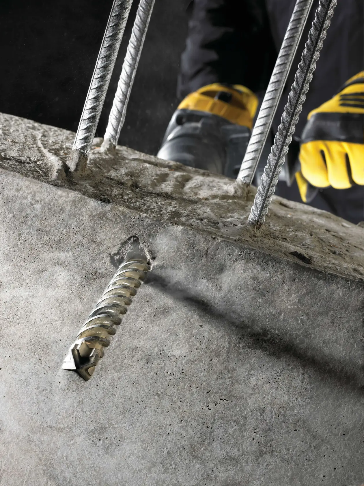 Broca DEWALT perforando concreto con barras de acero, usada por trabajador con guantes amarillos.