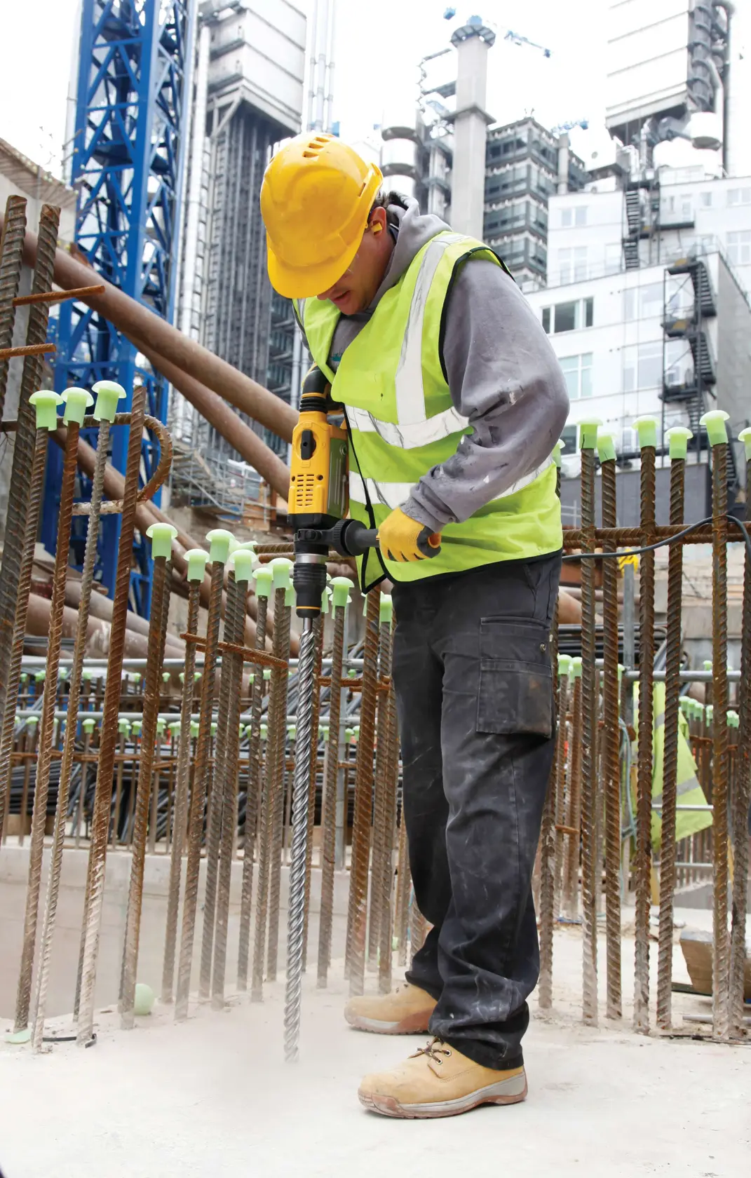 Arbeiter mit Schutzkleidung bohrt Beton mit DEWALT-Werkzeug auf Baustelle, umgeben von Bewehrungsstäben.