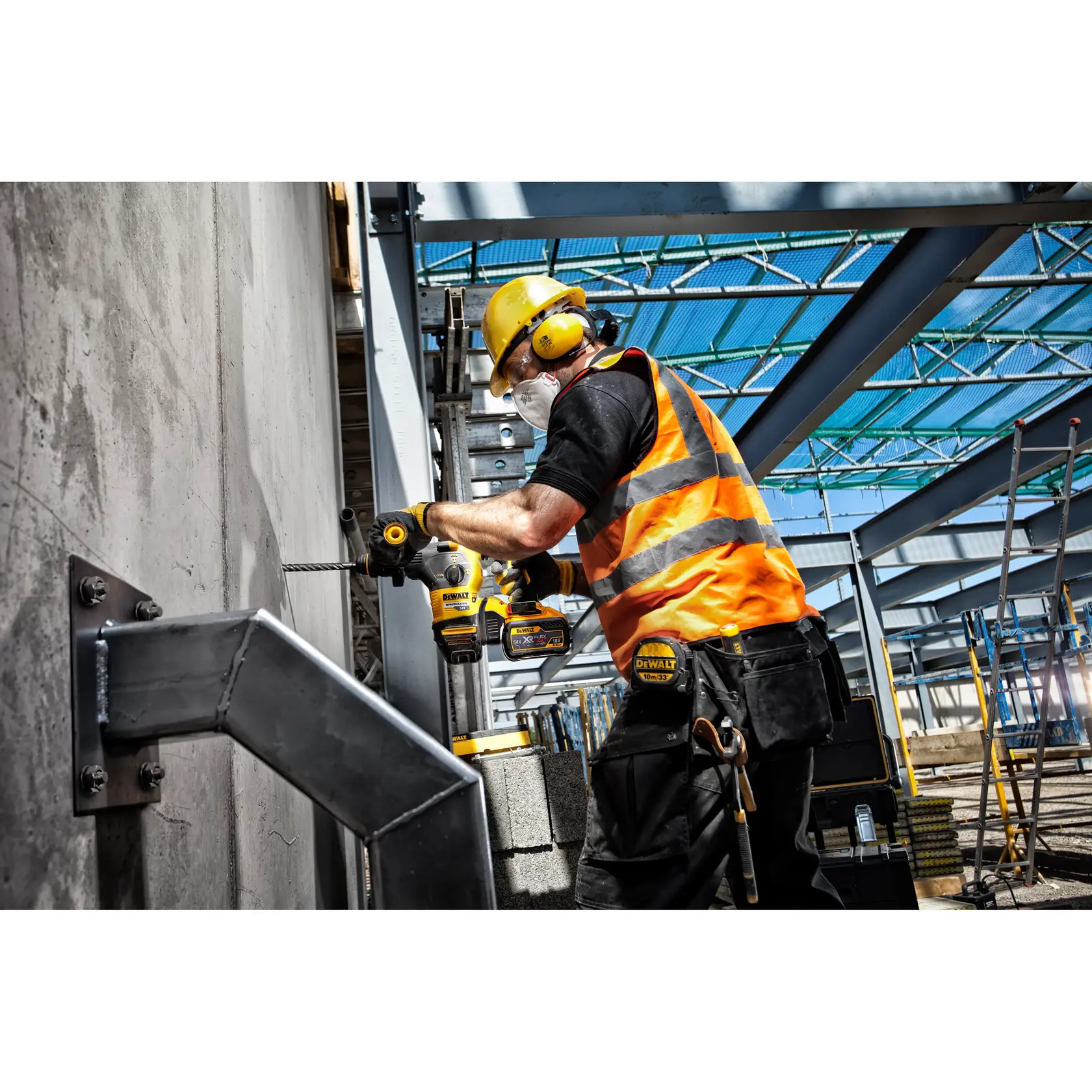 Un trabajador de la construcción con equipo de protección, que incluye casco amarillo, protección auditiva, gafas de seguridad y chaleco reflectante naranja, utiliza un rotomartillo inalámbrico DEWALT para perforar una pared de hormigón en una obra con vigas de acero y equipos al fondo.