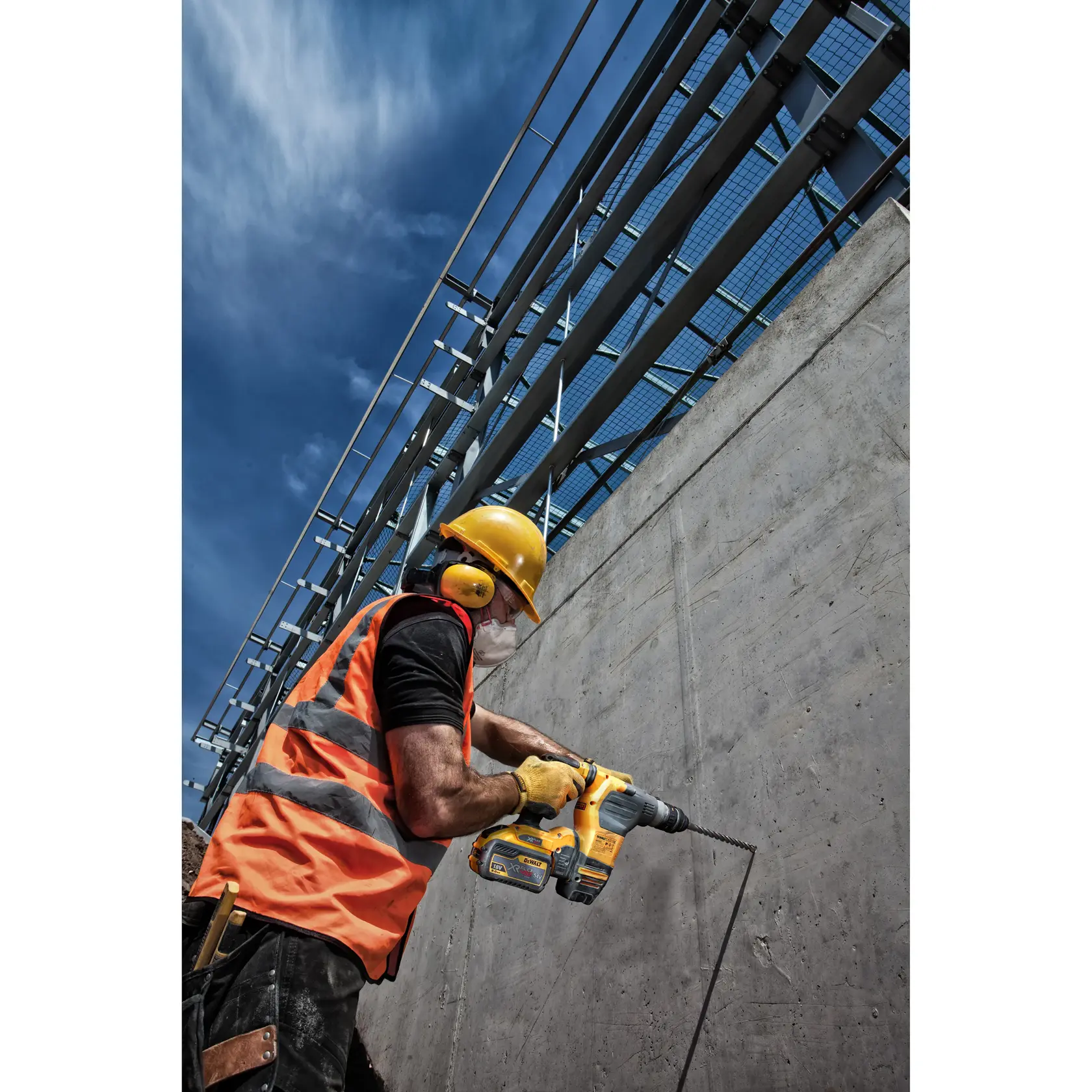 Trabajador con taladro amarillo y equipo de seguridad perfora pared de concreto en obra al aire libre.