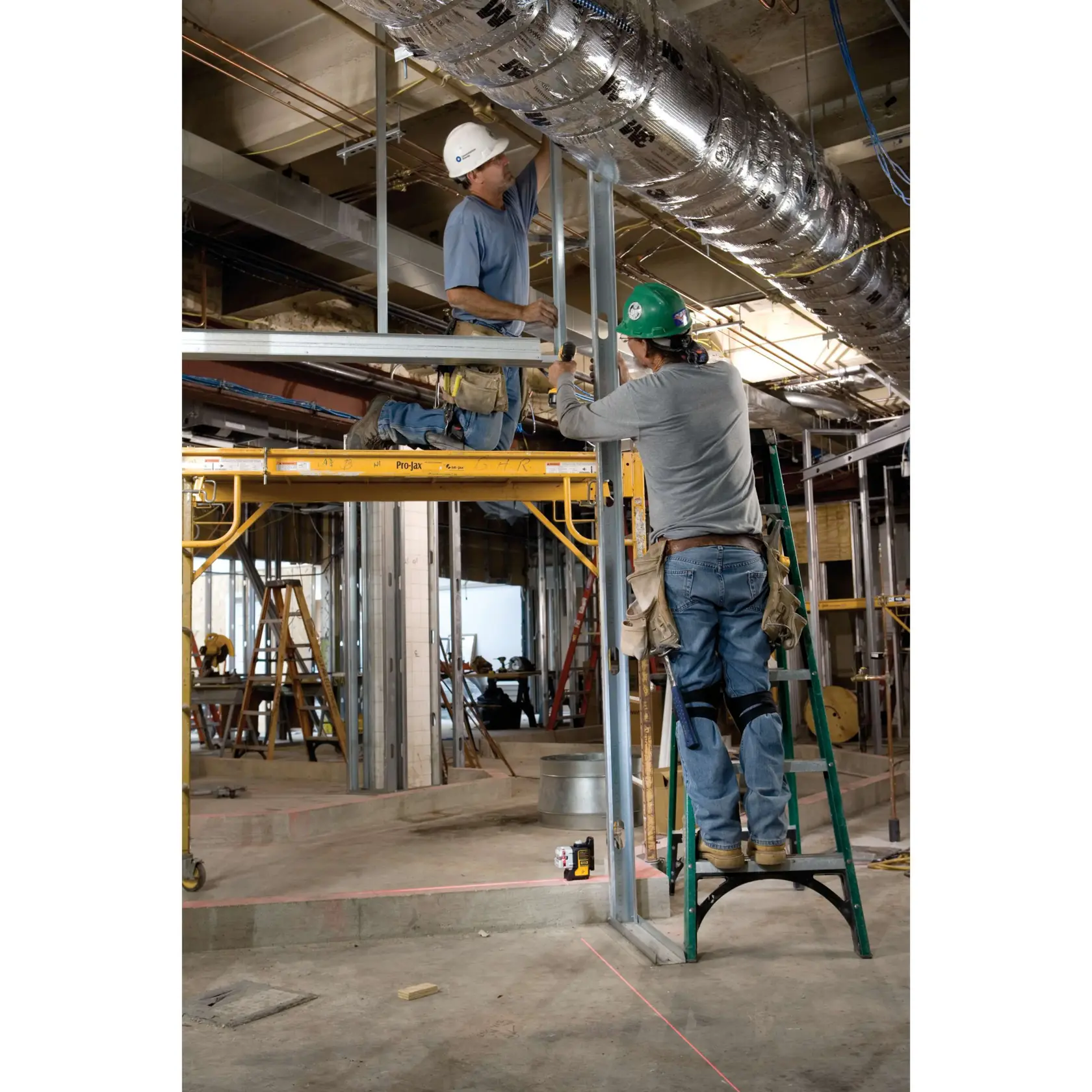 Un niveau laser DEWALT DW089K est placé au sol sur un chantier de construction, tandis qu’un ouvrier portant un casque et une ceinture à outils mesure avec un mètre ruban sur le sol. L’arrière-plan montre divers équipements de construction, des échelles et des tuyaux du plafond exposés.
