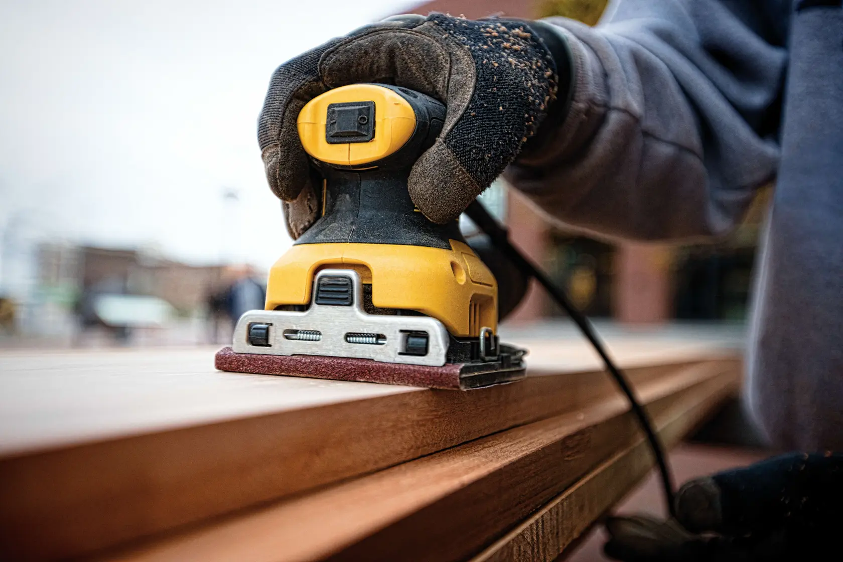 Eine Person verwendet einen DEWALT Elektroschleifer auf einer Holzoberfläche in einer Werkstatt oder Garage. Der Schleifer ist gelb und schwarz, und die Hand der Person hält ihn gegen das Holz.