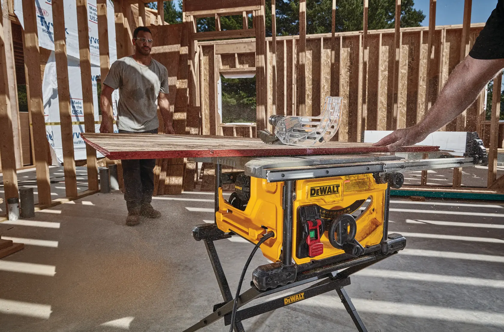 Eine gelbe DEWALT Tischsäge schneidet ein Holzstück in einem teilweise fertiggestellten Holzgebäude. Eine Person bedient die Säge, eine andere steht im Hintergrund. Sägemehl ist um die Maschine sichtbar.