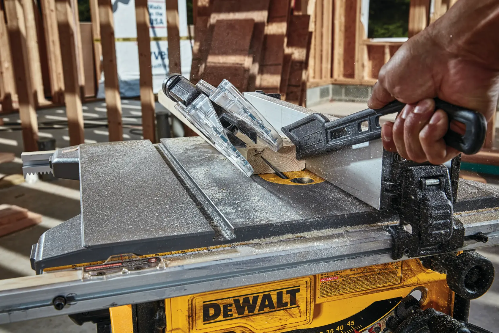 Nahaufnahme einer DEWALT Tischkreissäge im Einsatz, bei der eine Hand ein Holzstück mit einem Schiebestock durch das Sägeblatt führt. Die Säge wird auf einer Baustelle mit Holzrahmen im Hintergrund betrieben.